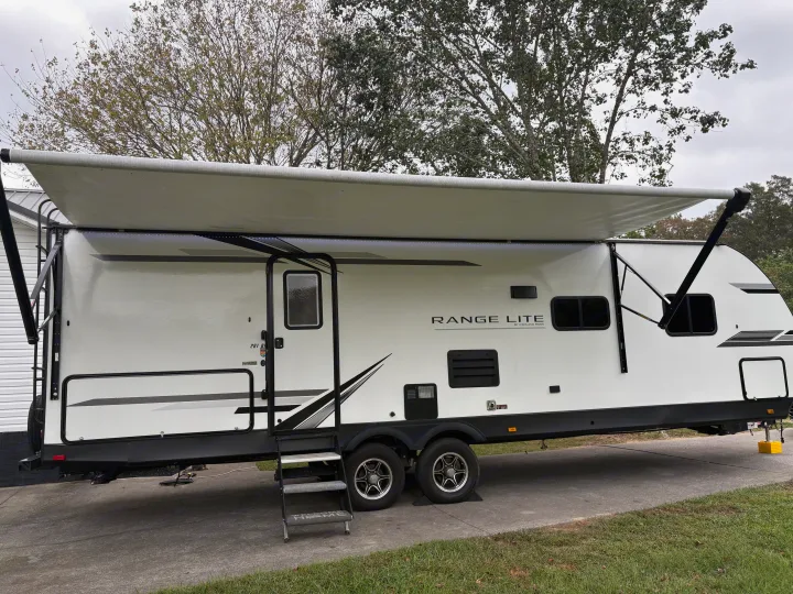 A white Range Lite travel trailer, ideal for an rv rental, is parked with its awning extended, framed by trees and concrete.. Highland Ridge RV Range Lite 2021