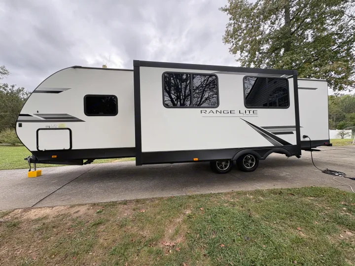 This white Range Lite travel trailer, an RV rental with its slide-out extended, is parked on a driveway with a power cable connected.. Highland Ridge RV Range Lite 2021