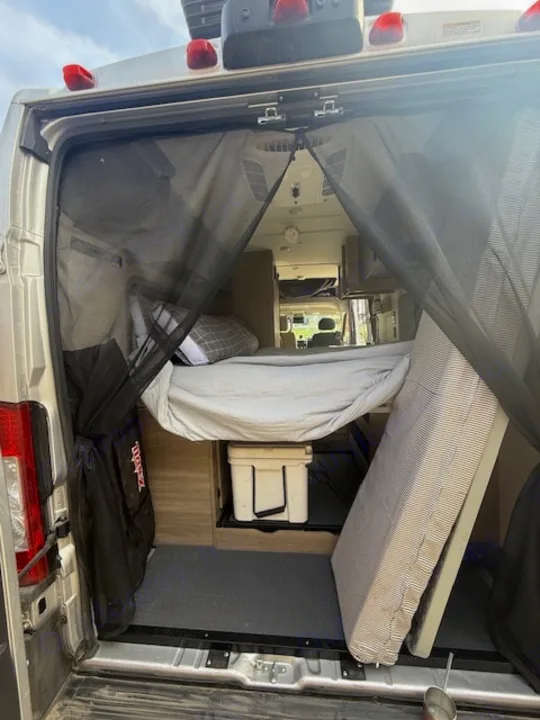 View into an RV rental from the open rear, revealing a mesh screen, raised bed, and interior storage including a cooler.. Winnebago Solis 2022