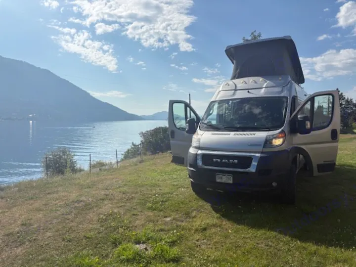 This RV rental camper van, with its pop-top extended, is parked on a grassy hill overlooking a scenic lake and mountains.. Winnebago Solis 2022