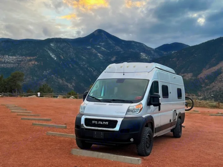 Experience mountain adventures! This silver camper van RV rental is parked on a reddish dirt lot, with snow-dusted mountains under a cloudy sky.. Winnebago Solis 2022