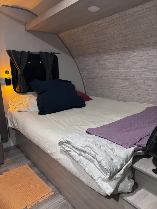 RV rental bedroom with a comfortable bed, dark blue & white pillows, blankets, and a side lamp by the window.. Coachmen 184BH 2025