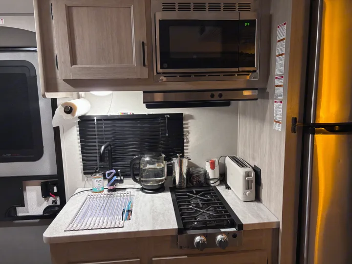 Inside an RV rental, this cozy kitchen features a microwave, gas cooktop, fridge, sink, and various small appliances.. Coachmen 184BH 2025
