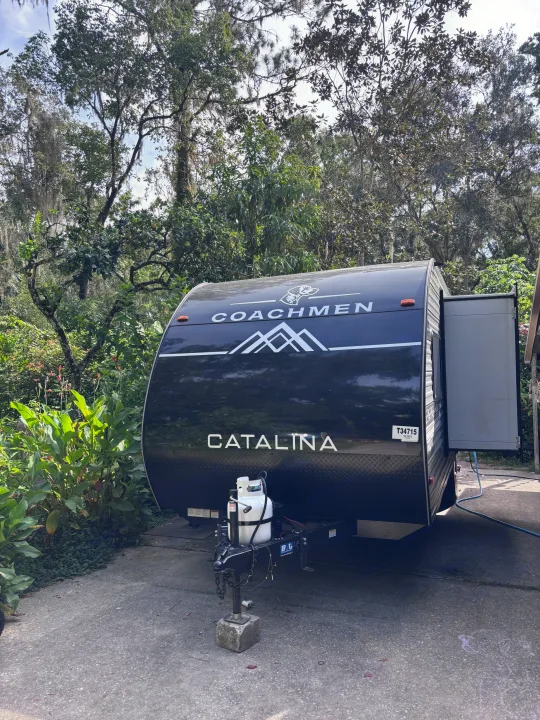 This black Coachmen Catalina travel trailer rv rental is parked on concrete amidst lush trees and greenery at a campsite.. Coachmen 184BH 2025