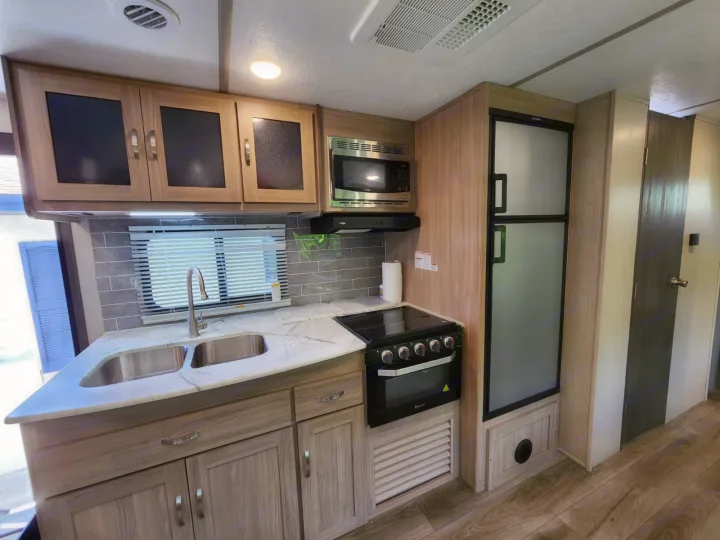 Modern RV rental kitchen featuring a double sink, stove, microwave, and fridge, all in light wood tones.. Coachmen 292BHDS 2021