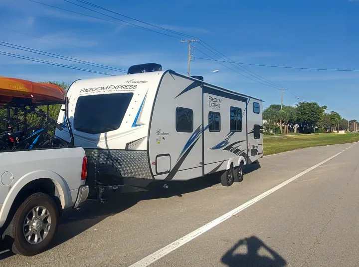 RV rental adventure begins! White pickup tows a Coachmen Freedom Express RV, loaded with kayaks and bikes, under a blue sky.. Coachmen 292BHDS 2021