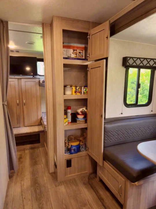 View inside an RV rental with an open, well-stocked pantry, a dining booth, and a TV area beyond.. Coachmen 292BHDS 2021