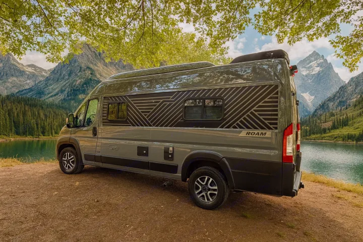 This RV rental is parked by a serene mountain lake, surrounded by lush forest and majestic peaks, ideal for an outdoor adventure.. Winnebago Roam 2024