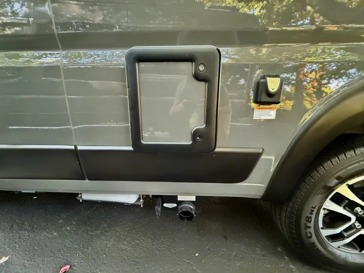 Side of an RV rental, showing a utility access panel, electrical inlet, and gray water tank drain valve for waste disposal.. Winnebago Roam 2024