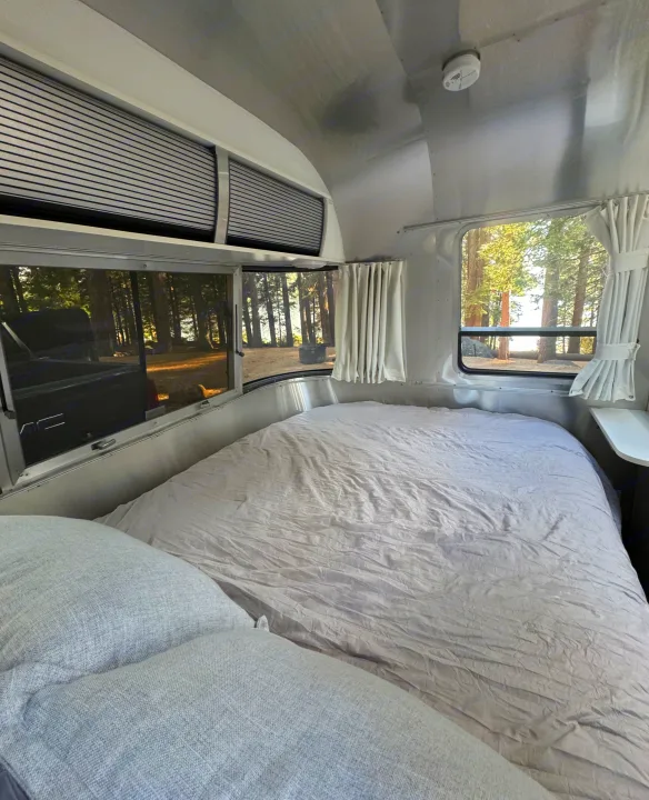 Cozy Airstream RV rental bedroom: light bedding, pillows, and large windows revealing a forest and distant water.. Airstream Sport 2018