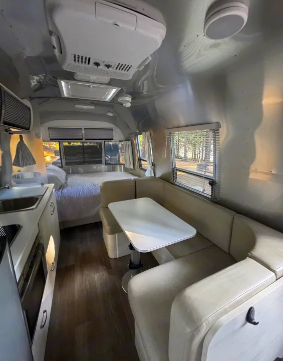 This rv rental features a sleek, silver interior with a cozy bed, compact kitchen, and inviting U-shaped dining area.. Airstream Sport 2018