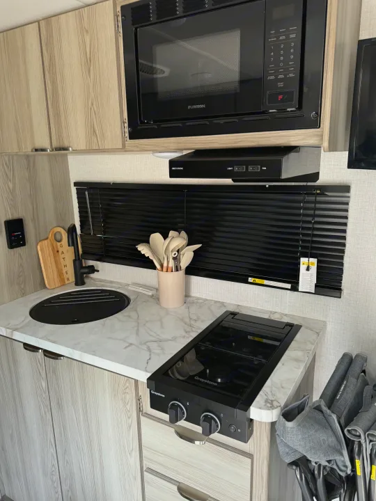 This rv rental kitchen features a compact design with a microwave, sink, Greystone stove, and modern light wood cabinets.. Forest River Salem 2024