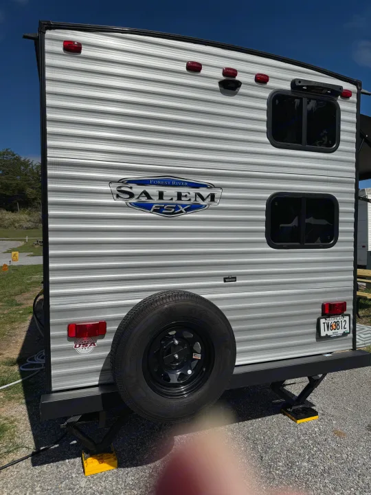 Silver Forest River Salem FSX RV rental's rear, featuring spare tire, taillights, and stabilizers on gravel, ready for camping.. Forest River Salem 2024