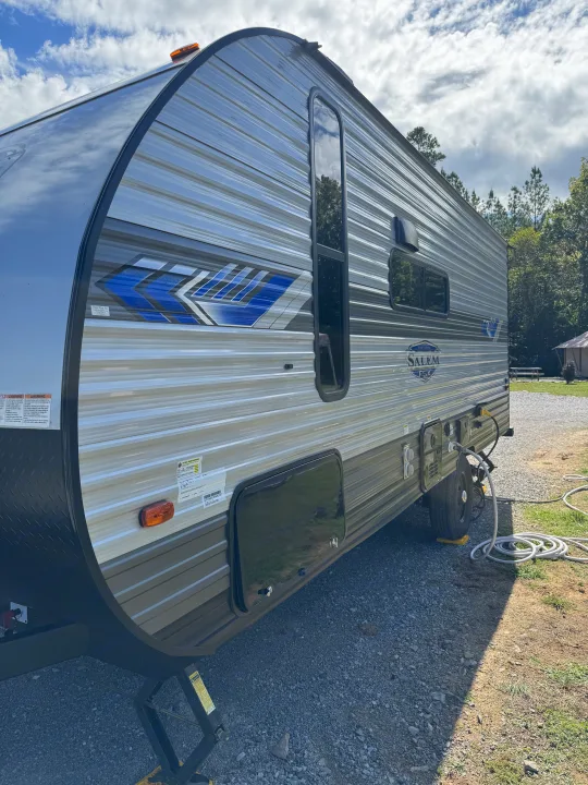 A modern silver and blue Salem FSX RV rental is parked on a gravel campsite, hooked up for an outdoor adventure.. Forest River Salem 2024