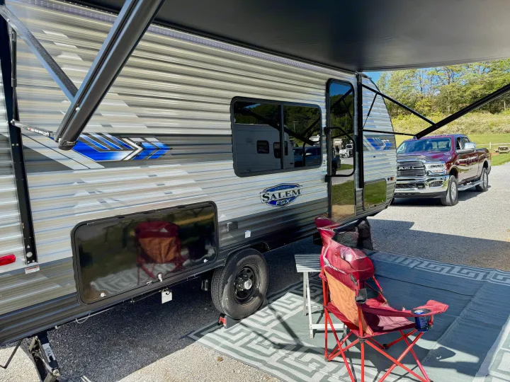 Sunny RV rental: A modern travel trailer, with its awning extended over an outdoor rug and chair, alongside a red pickup truck.. Forest River Salem 2024