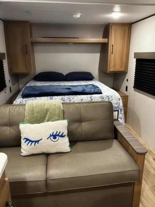 Cozy RV rental interior with a bed (blue/patterned bedding), a gray couch with an eye pillow, and light wood overhead storage.. Coleman 23B Keystone 2025