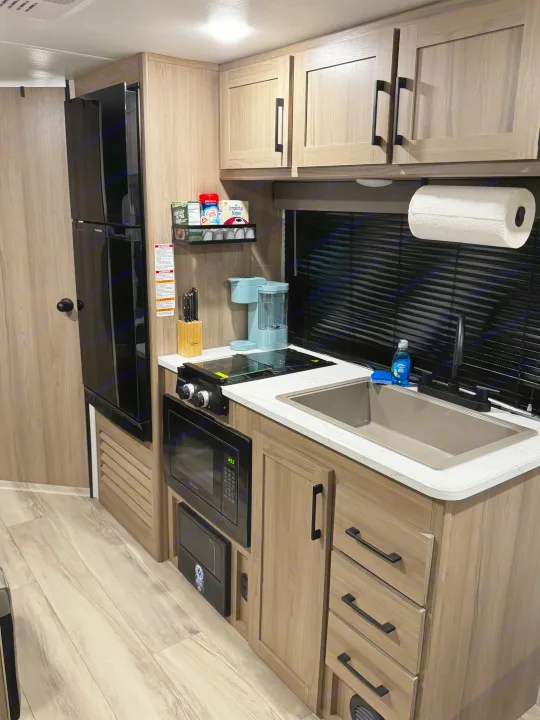 A clean, modern rv rental kitchen featuring light wood cabinets, black appliances, a coffee maker, and a compact sink.. Coleman 23B Keystone 2025