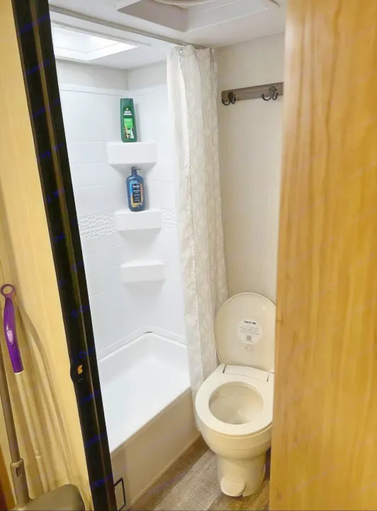 Compact rv rental bathroom with a shower (shampoo on shelves), toilet (lid up), and wall hooks. A purple mop is on the left.. Forest River Campsite reserve 25MW 2024