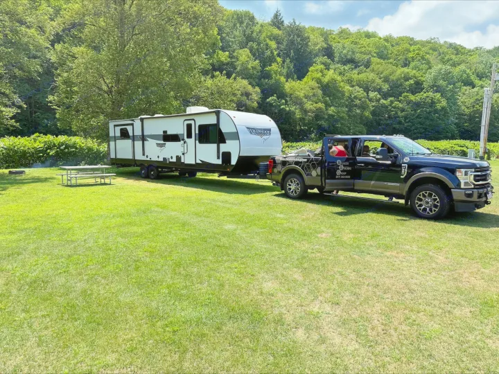 A black truck is towing a white rv rental on a sunny, grassy campsite. Trees and a picnic table are in the background.. Forest River Wildwood 2024