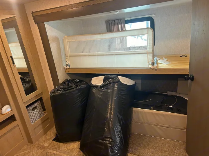 Inside an RV rental: a bunk bed with a child safety rail, storage shelves, and large black bags, possibly for moving or cleanup.. Forest River Wildwood 2024
