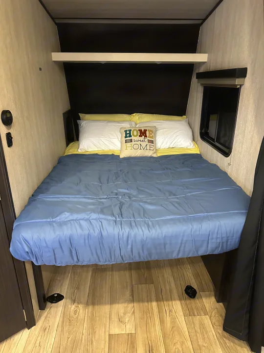 An RV rental bedroom features a made-up bed with a blue comforter and "HOME sweet HOME" pillow, offering a compact sleeping area.. Genesis Supreme Rv Genesis Supreme 2025