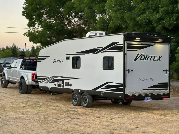 A large silver dually truck tows a white "Vortex" toy hauler on dry grass. Perfect for an RV rental seeking adventure.. Genesis Supreme Rv Genesis Supreme 2025