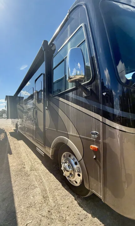 Explore in this brown RV rental, a large motorhome with an awning, parked on a dirt lot under blue skies.. Coachmen Encore 2022