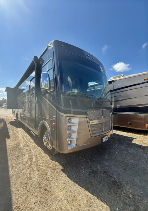 A dark, large Class A RV rental is parked on a dirt lot with an extended awning. Clear blue sky above, another RV partly visible.. Coachmen Encore 2022
