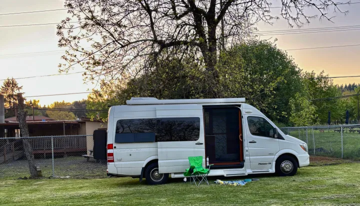 A white Pleasure-Way camper van, a perfect RV rental, parked on a grassy lawn at sunset with a chair and blanket for outdoor leisure.. Pleasure-Way Pleasure-Way Class B 2016