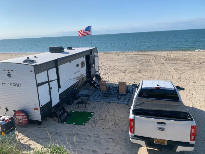 An RV rental and pickup truck are beach camping by the ocean. An American flag flies over the scenic setup, perfect for outdoor relaxation.. Keystone RV Hideout 2021