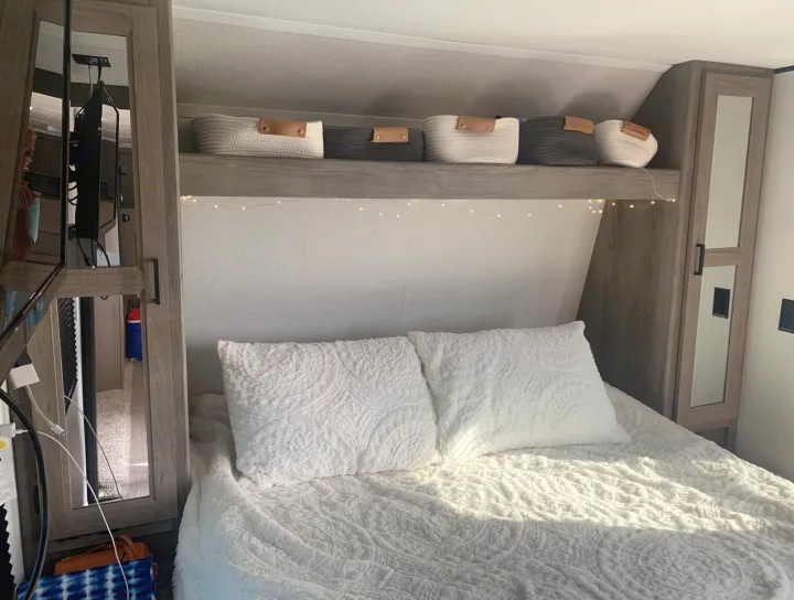 Cozy RV rental bedroom interior with a comfy white bed, woven baskets on a shelf, and warm fairy lights.. Keystone RV Hideout 2021