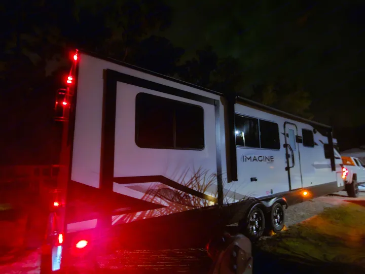 A white Imagine **rv rental** travel trailer is parked at night, its red tail lights casting a prominent glow, with a truck visible behind it.. Grand Design Imagine 2024