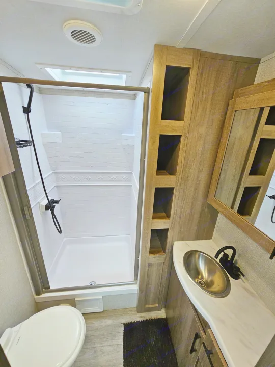 Compact RV rental bathroom: walk-in shower, toilet, modern vanity with stainless steel sink, black fixtures, and wood shelving.. Grand Design Imagine 2024