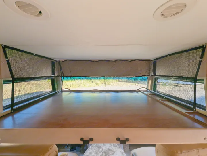 Inside an RV rental: a cozy overhead bunk with safety netting, visible windows showing trees, and ceiling vents for comfort.. Thor Miramar 2015