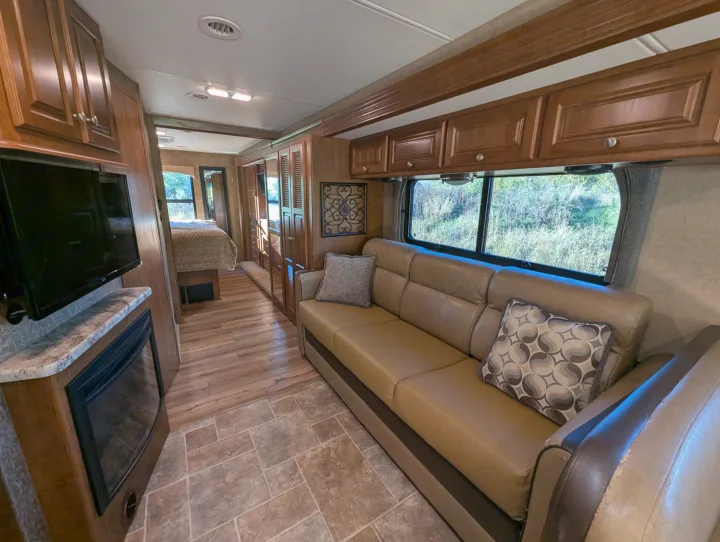 This RV rental features a living area with a tan leather sofa, TV, fireplace, and wood-look flooring leading to a bedroom.. Thor Miramar 2015