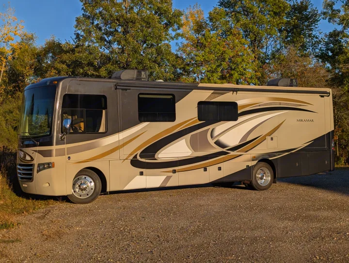 A sleek, two-tone Miramar RV rental is parked on a gravel lot with trees and a blue sky in the background.. Thor Miramar 2015