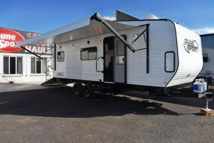 Modern white toy hauler RV rental with awning extended & door open, parked on asphalt. Propane tanks are visible.. Dune Sport Dynamite 2023