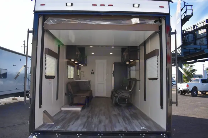 Inside a modern RV rental toy hauler, viewed from the open rear ramp. Features a kitchen, fold-down sofa, and lounge chairs.. Dune Sport Dynamite 2023