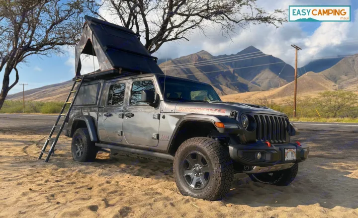 Gray Jeep Gladiator RV rental with open rooftop tent & ladder on sand, mountains in background. Ideal for Maui camping.. Jeep Gladiator 2022