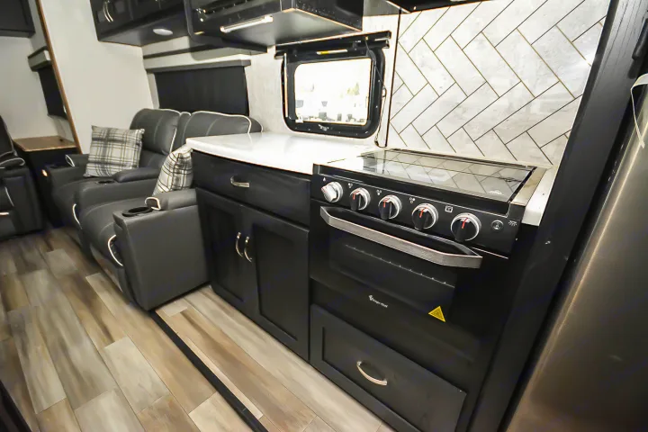 Inside a modern RV rental: sleek kitchen with black oven, white counter, herringbone tile, next to comfy gray recliners.. Coachmen Coachmen Freedom Express Liberty Edition (324RLDSLE) 2022