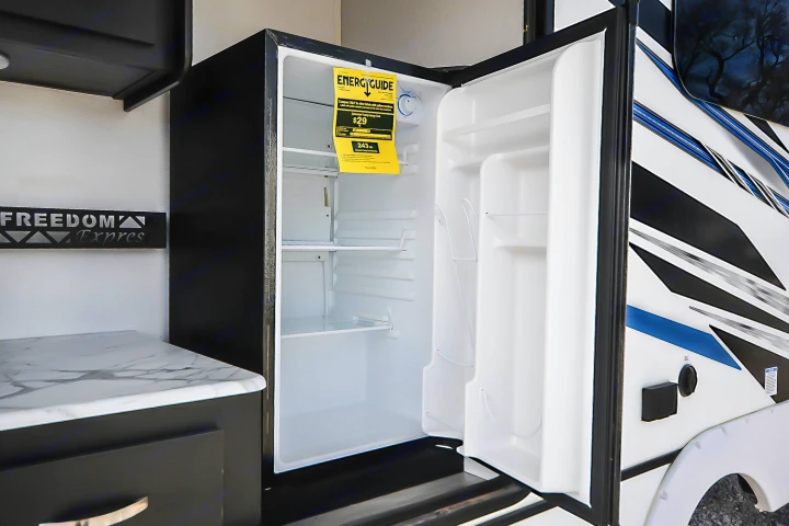 Open, compact mini-fridge inside an RV rental, with "Freedom Express" branding and a counter on the left.. Coachmen Coachmen Freedom Express Liberty Edition (324RLDSLE) 2022