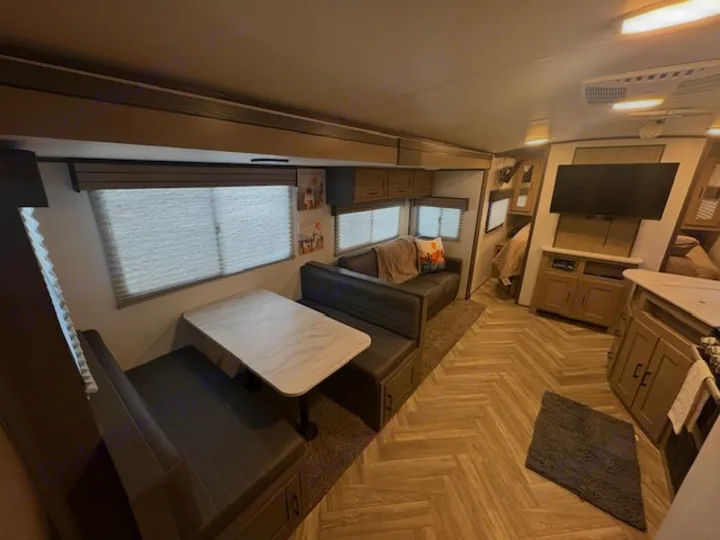 An RV rental interior shows a dinette with benches & table, a sofa, kitchen island, and TV. Light-colored herringbone floor.. Cruiser RV Shadow Cruiser 2021