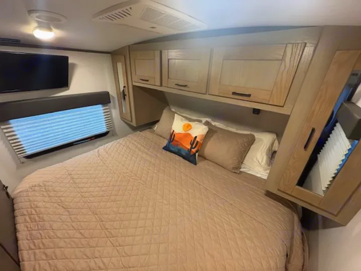 Cozy RV rental bedroom featuring a large bed, wall-mounted TV, window with blinds, and ample storage overhead.. Cruiser RV Shadow Cruiser 2021