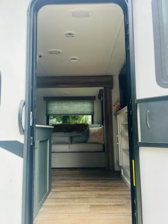 Open RV rental door reveals a cozy living space with sofa, window, wood-look flooring, and built-in cabinets.. Heartland RVs 265BH-L 2024