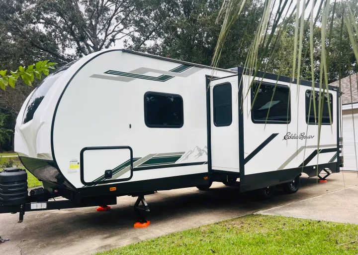 An Eddie Bauer travel trailer, white with green/grey graphics, slide-out extended, parked on a driveway. Ideal for rv rental adventures!. Heartland RVs 265BH-L 2024