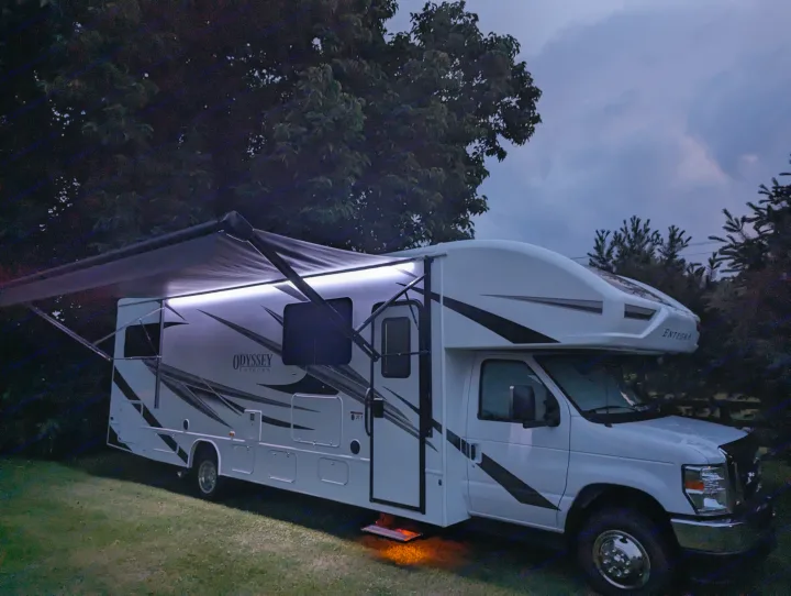 White Class C rv rental with glowing awning and step lights at dusk, parked on grass against a dark, cloudy sky.. Entegra Coach Odyssey 2024