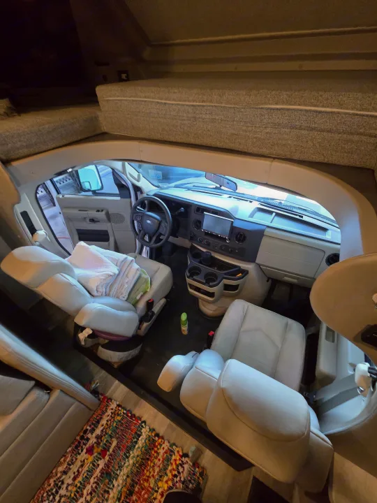 Spacious RV rental interior featuring the cockpit, dashboard, light-colored seats with towels, and an overhead bunk.. Entegra Coach Odyssey 2024