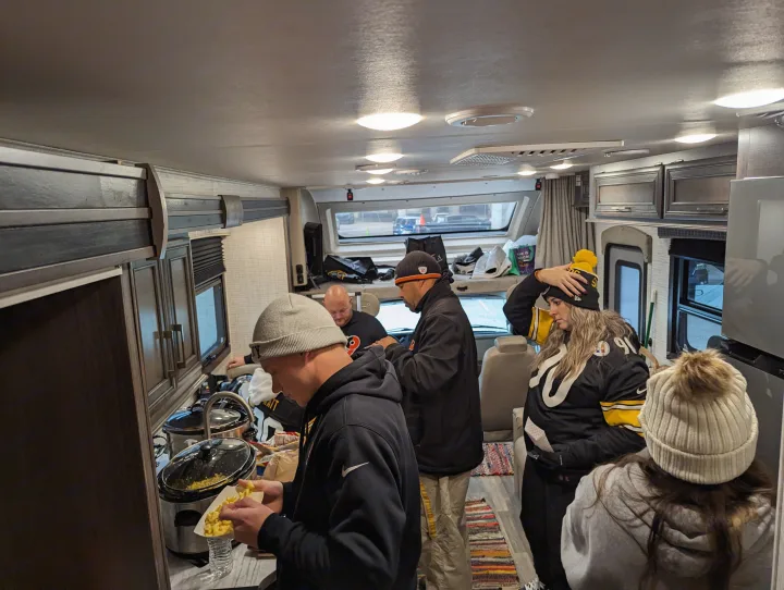 Friends enjoying mac & cheese in an RV rental, dressed in team gear. Perfect for a tailgate party!. Entegra Coach Odyssey 2024