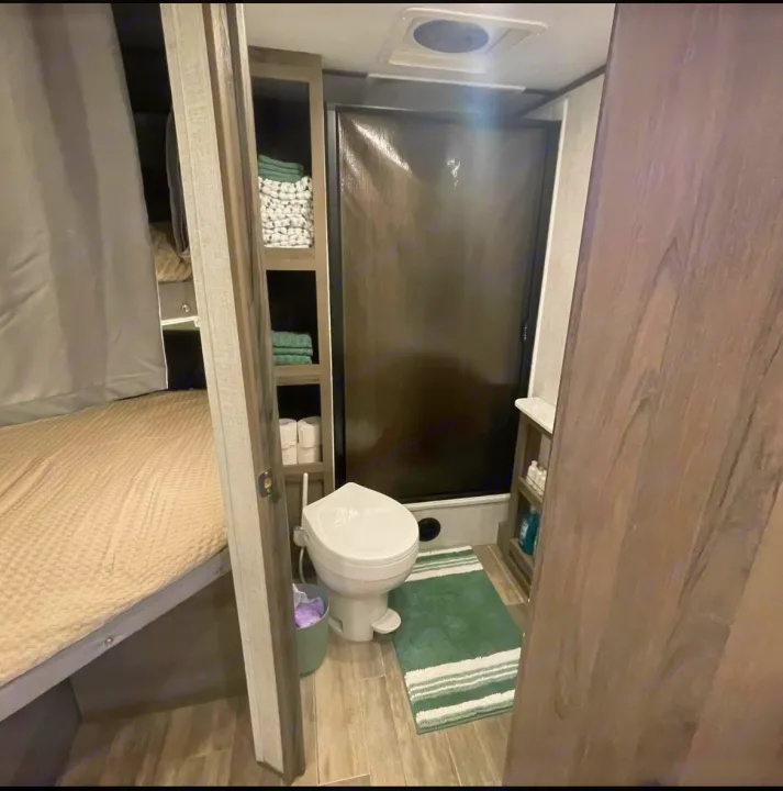 This compact RV rental bathroom includes a toilet, shower, storage shelves with towels, and a peek at the adjacent bed area.. Gulf Stream Envision 2023