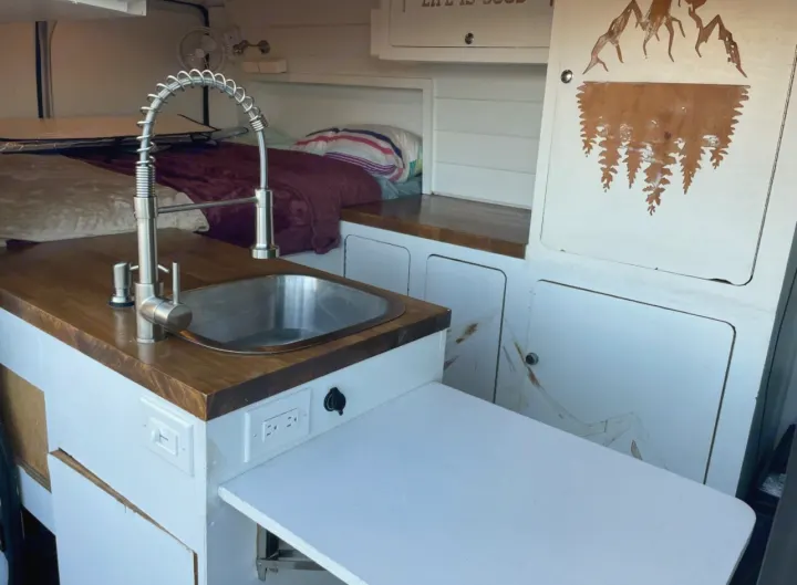 Cozy RV rental interior featuring a kitchen sink, pull-out counter, outlets, and a plush sleeping area in the background.. Ram Promaster 2021